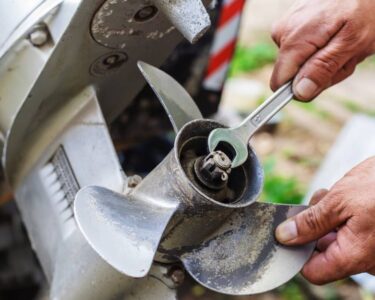 A boat's dirty outboard motor propeller. Two hands are holding a wrench and the propeller to loosen nuts and bolts.