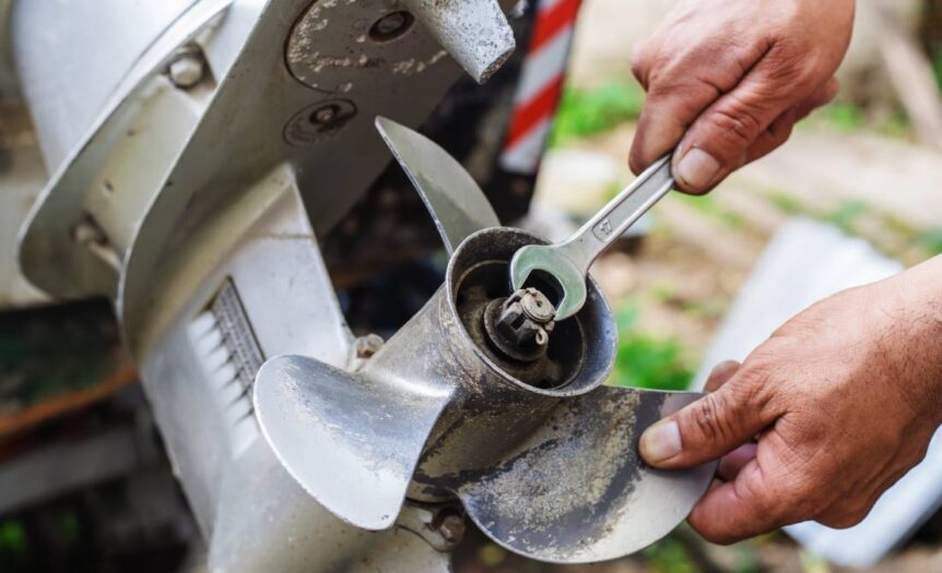 A boat's dirty outboard motor propeller. Two hands are holding a wrench and the propeller to loosen nuts and bolts.