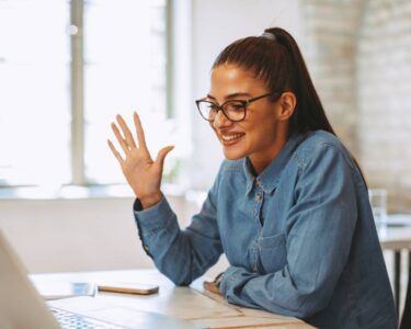 A young woman in a blue top leaning against her desk and waving at her laptop during a teleconference interview.