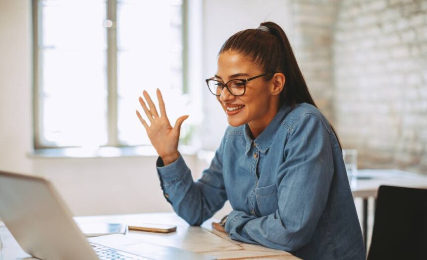 A young woman in a blue top leaning against her desk and waving at her laptop during a teleconference interview.