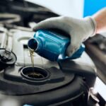 A close-up of a gloved hand holding a blue container and pouring motor oil into the engine compartment of a vehicle.