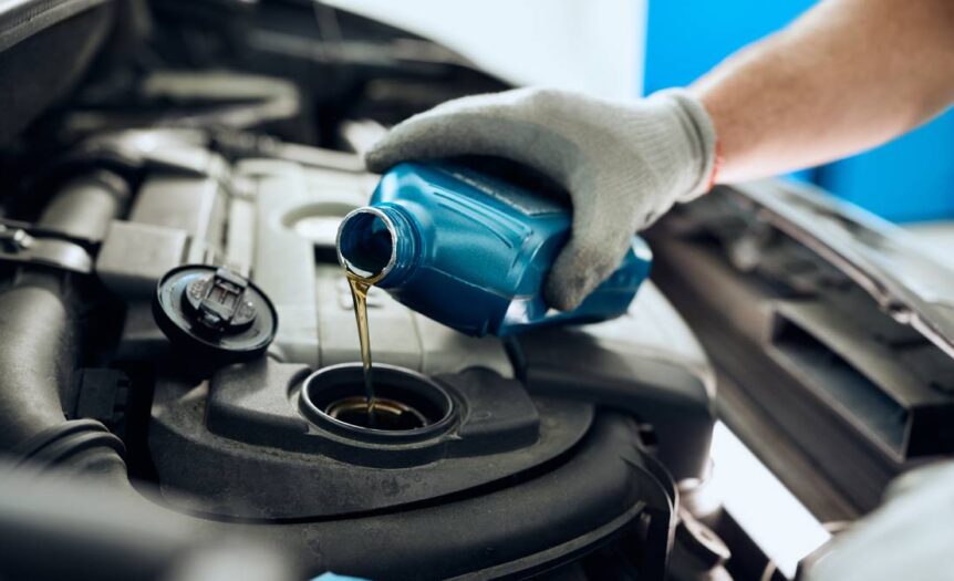 A close-up of a gloved hand holding a blue container and pouring motor oil into the engine compartment of a vehicle.