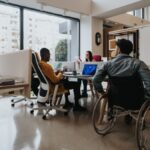 An office with ergonomic furniture and multicultural employees. A wheelchair user is near a shared employee desk.