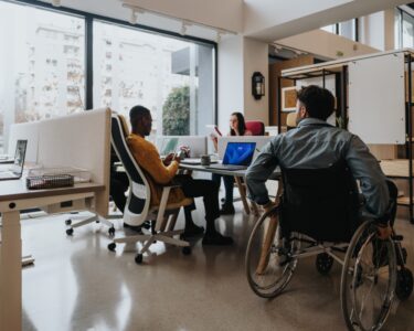 An office with ergonomic furniture and multicultural employees. A wheelchair user is near a shared employee desk.