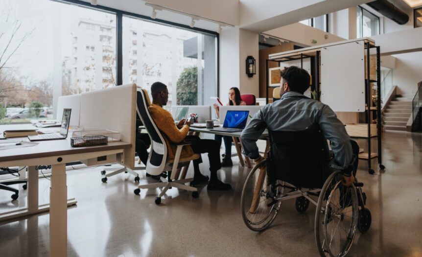 An office with ergonomic furniture and multicultural employees. A wheelchair user is near a shared employee desk.