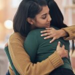 A woman wearing a brown sweater wrapping both arms around a person. She has her eyes shut and a relaxed expression.