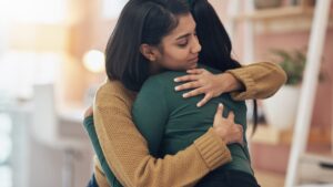 A woman wearing a brown sweater wrapping both arms around a person. She has her eyes shut and a relaxed expression.