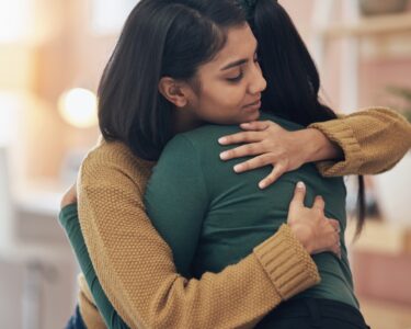 A woman wearing a brown sweater wrapping both arms around a person. She has her eyes shut and a relaxed expression.