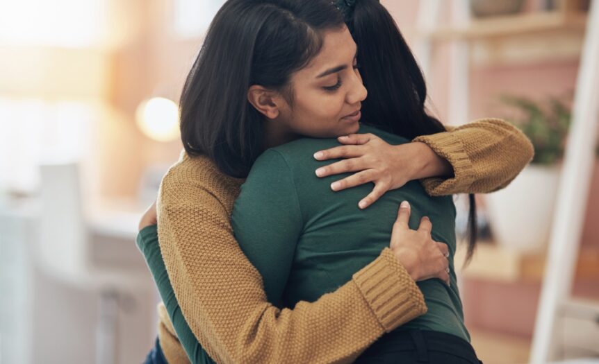 A woman wearing a brown sweater wrapping both arms around a person. She has her eyes shut and a relaxed expression.