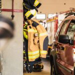 Fire-resistant clothing hanging on a hook inside of a firehouse. There are firefighter vehicles next to the clothes.