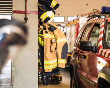 Fire-resistant clothing hanging on a hook inside of a firehouse. There are firefighter vehicles next to the clothes.