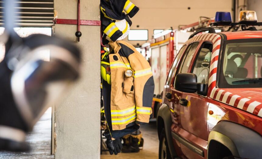 Fire-resistant clothing hanging on a hook inside of a firehouse. There are firefighter vehicles next to the clothes.