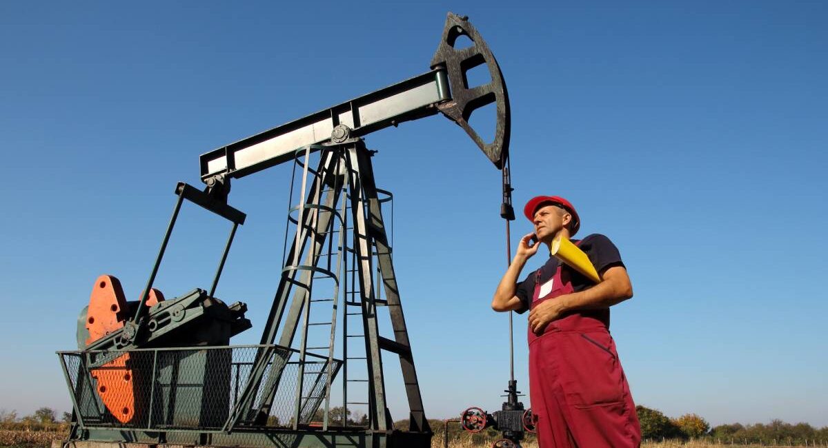 A man in red overalls and a red construction hat stands in front of an oil rig. He's talking on the phone and holding papers.