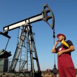 A man in red overalls and a red construction hat stands in front of an oil rig. He's talking on the phone and holding papers.