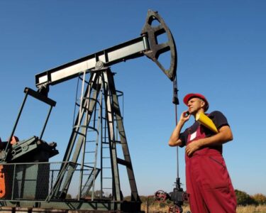 A man in red overalls and a red construction hat stands in front of an oil rig. He's talking on the phone and holding papers.