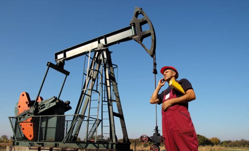 A man in red overalls and a red construction hat stands in front of an oil rig. He's talking on the phone and holding papers.