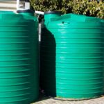 Two green plastic water tanks next to a property. There are trees, a hose and potted plants next to the tanks.