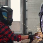 A female mechanic wearing safety goggles and welding gloves, concentrating as bright sparks fly during the welding process.