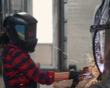 A female mechanic wearing safety goggles and welding gloves, concentrating as bright sparks fly during the welding process.