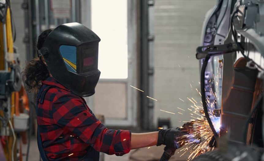 A female mechanic wearing safety goggles and welding gloves, concentrating as bright sparks fly during the welding process.