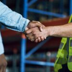 Close-up of two men shaking hands. One man is wearing a long-sleeved shirt, and the other is wearing a high-visibility vest.