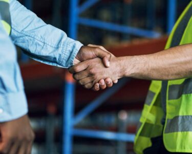 Close-up of two men shaking hands. One man is wearing a long-sleeved shirt, and the other is wearing a high-visibility vest.