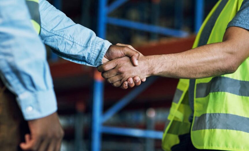 Close-up of two men shaking hands. One man is wearing a long-sleeved shirt, and the other is wearing a high-visibility vest.