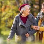 A young woman in a yellow coat is walking in a park with an older woman in a grey coat and burgundy hat.