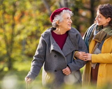 A young woman in a yellow coat is walking in a park with an older woman in a grey coat and burgundy hat.