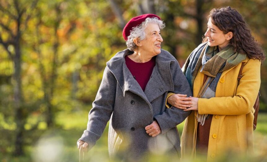 A young woman in a yellow coat is walking in a park with an older woman in a grey coat and burgundy hat.