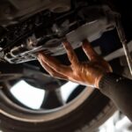 A person working on their car lets transmission fluid drain out of the bottom of the vehicle. A wheel is in the background.