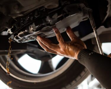 A person working on their car lets transmission fluid drain out of the bottom of the vehicle. A wheel is in the background.