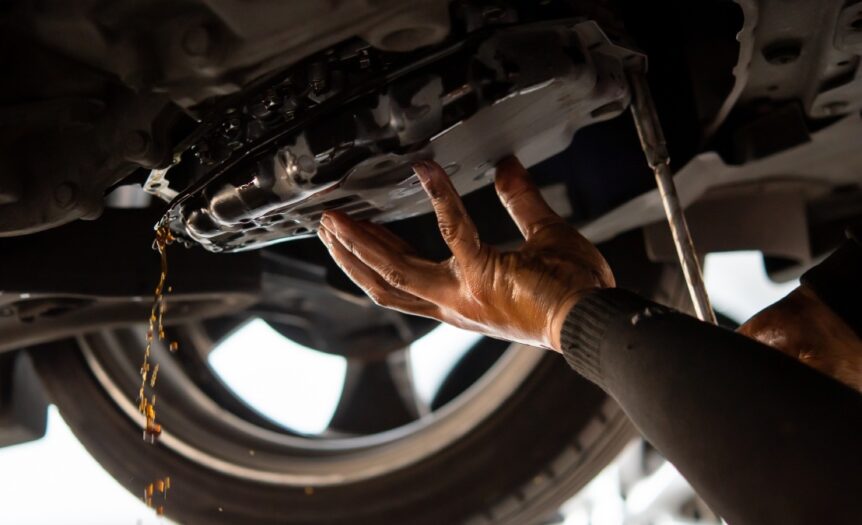 A person working on their car lets transmission fluid drain out of the bottom of the vehicle. A wheel is in the background.