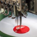 A machine embroidery needle during operation sewing a red and white design into a white fabric held by a stabilizer.