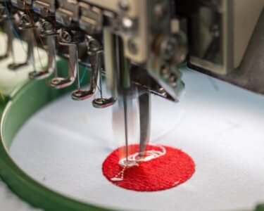 A machine embroidery needle during operation sewing a red and white design into a white fabric held by a stabilizer.