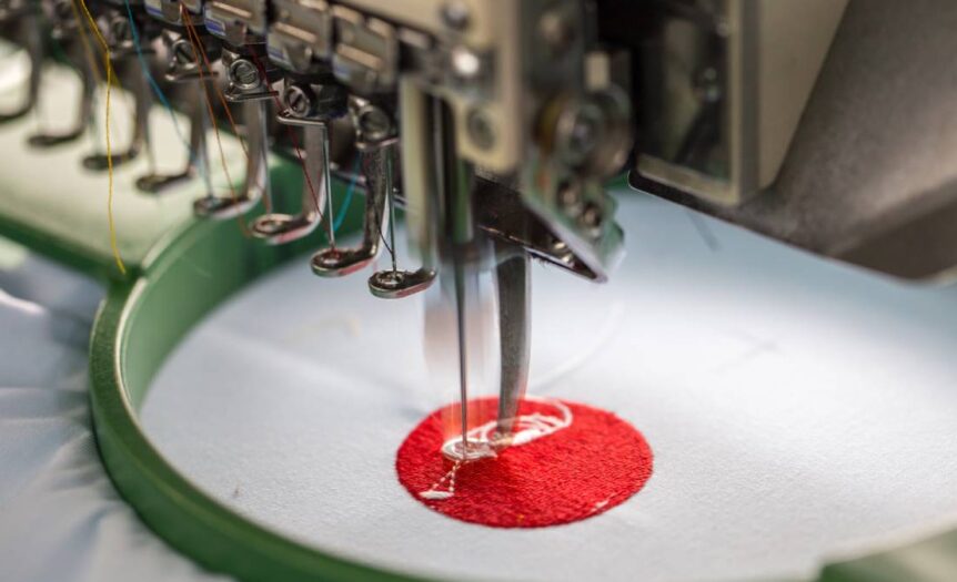 A machine embroidery needle during operation sewing a red and white design into a white fabric held by a stabilizer.