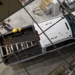 A person is talking on their phone while standing next to a pickup truck that is holding a ladder in the truck bed.