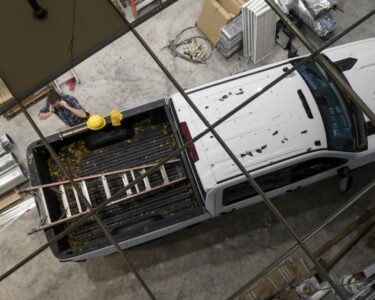 A person is talking on their phone while standing next to a pickup truck that is holding a ladder in the truck bed.