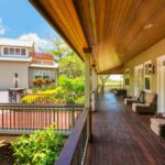 Side view of a long, luxurious front porch with dark hardwood floors, plush outdoor furniture, and surrounded by plants.