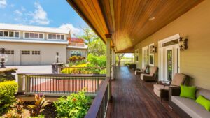 Side view of a long, luxurious front porch with dark hardwood floors, plush outdoor furniture, and surrounded by plants.