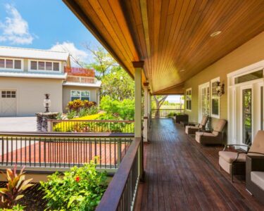 Side view of a long, luxurious front porch with dark hardwood floors, plush outdoor furniture, and surrounded by plants.