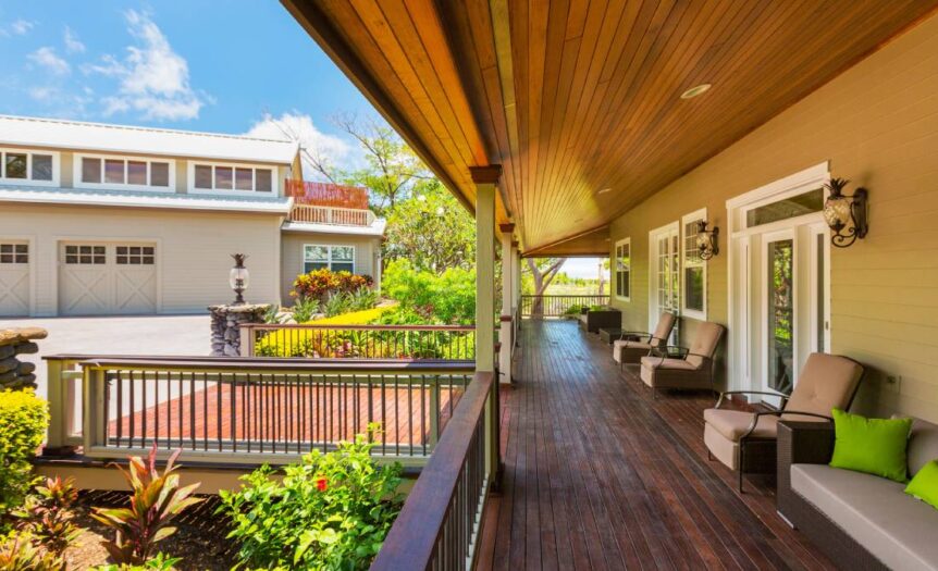 Side view of a long, luxurious front porch with dark hardwood floors, plush outdoor furniture, and surrounded by plants.