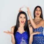 Two young women in formal blue dresses stand close to one another. One woman holds a tierra above the other’s head.