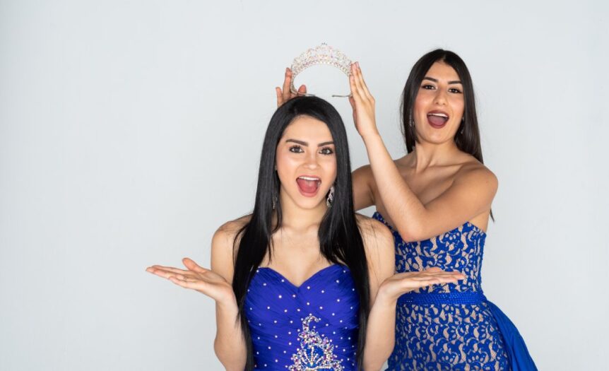 Two young women in formal blue dresses stand close to one another. One woman holds a tierra above the other’s head.