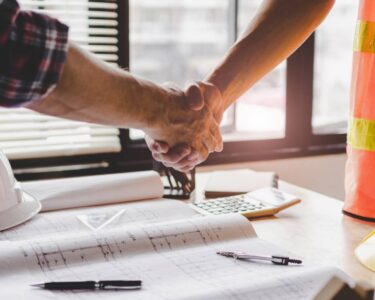 Two men shake hands over a desk with a hard hat and plans. One wears a flannel shirt, and the other wears a safety vest.