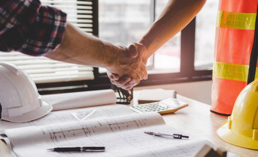 Two men shake hands over a desk with a hard hat and plans. One wears a flannel shirt, and the other wears a safety vest.