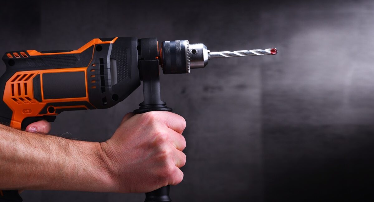 Two hands hold tightly onto an orange and black power drill. A gray background stands behind the drill.