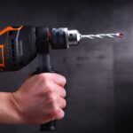 Two hands hold tightly onto an orange and black power drill. A gray background stands behind the drill.