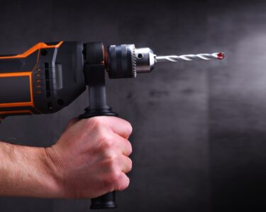 Two hands hold tightly onto an orange and black power drill. A gray background stands behind the drill.