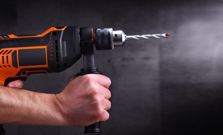 Two hands hold tightly onto an orange and black power drill. A gray background stands behind the drill.